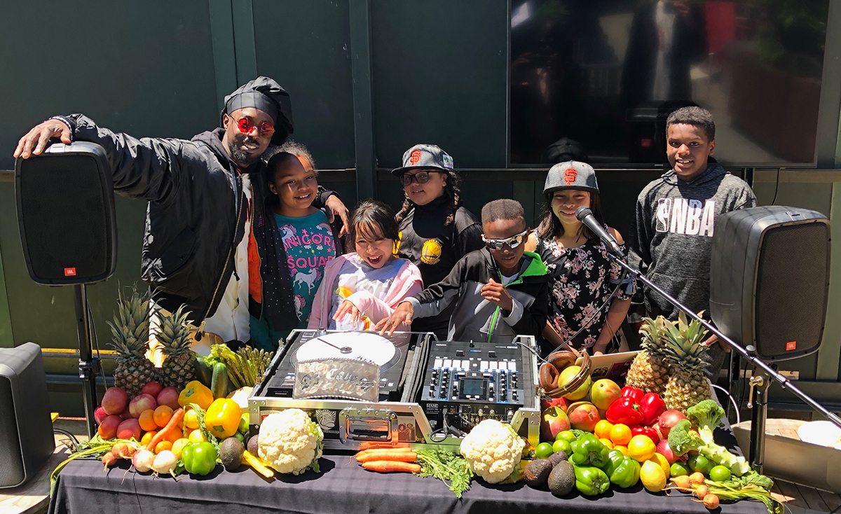 DJ Cavem poses behind his produce-covered DJ booth with students from Enchantment Institute