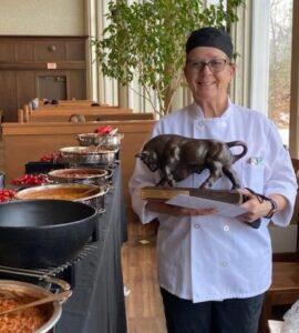 Female chef and winner of chili contest stands next to bowls of chili while holding a trophy