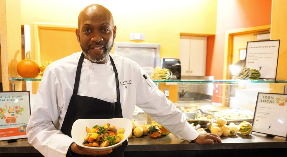 Smiling chef holds bowl of slow-roasted sweet maple-barbecue chili brisket with herb-roasted Yukon gold potatoes and corn on the cob in warmly lit room