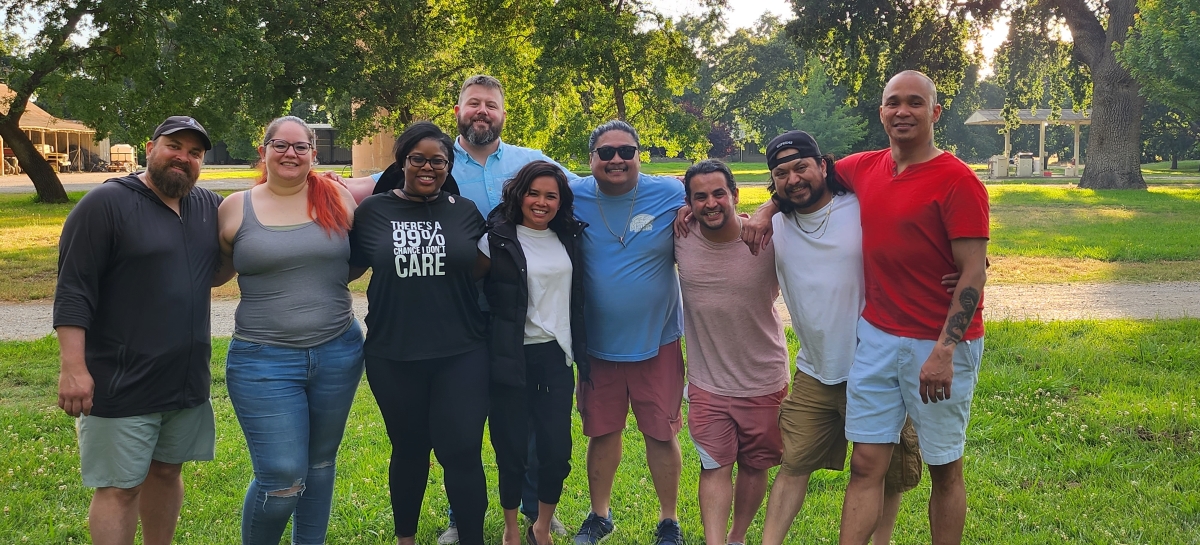 A group of people smiling and posing together under trees and dappled sunlight