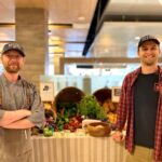 Two men stand in front of a display of produce.