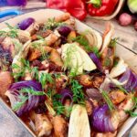 A dish of roasted chicken and onions with fresh produce in the background.
