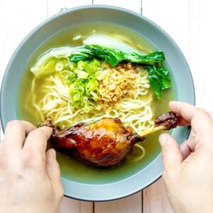 An image of a person placing a duck leg into a bowl of noodle soup.