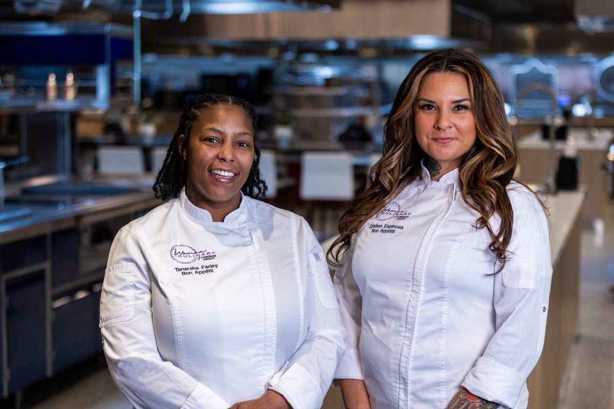 Two women in culinary whites stand together in a professional kitchen. 