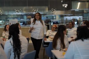A woman in a white chef's coat stands and addresses a group of other women chefs seated around her. 