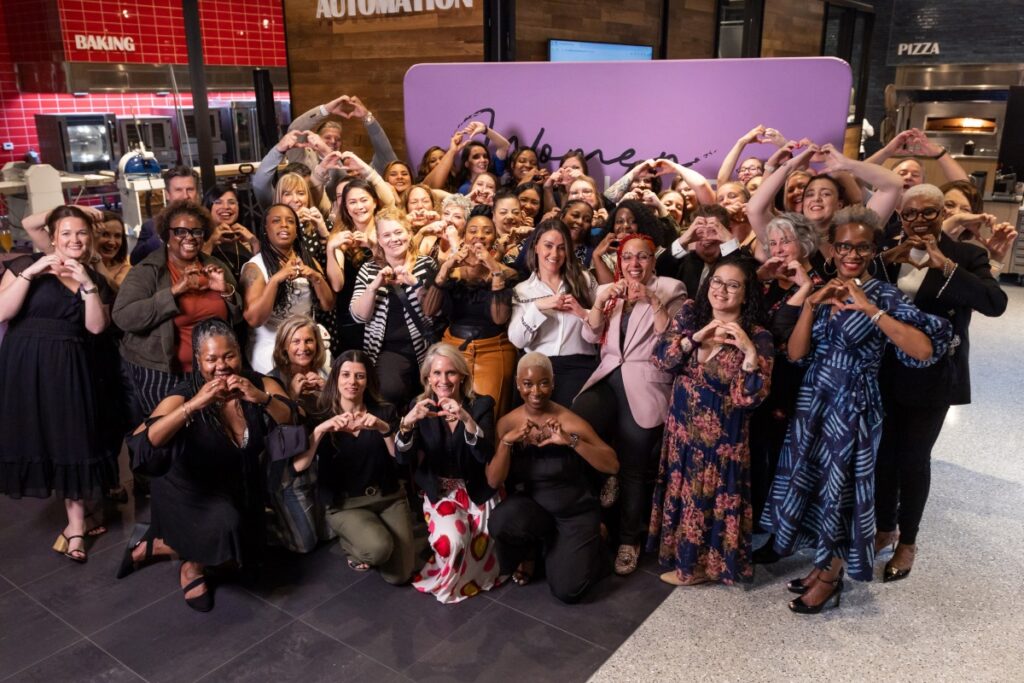 A large group of women gather in front of a purple sign and make heart hand gestures.