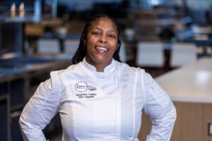 A woman in a white chef's coat stands smiling in a commercial kitchen