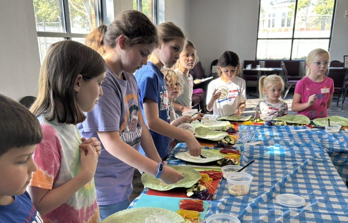 A group of children attend a cooking demonstration with brightly colored placcemats and tablecloths under their fruits and vegetables.