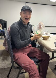 A man is seated at a table with a big smile on his face, holding a bag of ingredients for ice cream