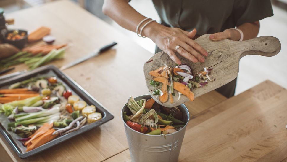 A woman's arm and hand is shown moving produces scraps from a cutting board to a compost bin. 