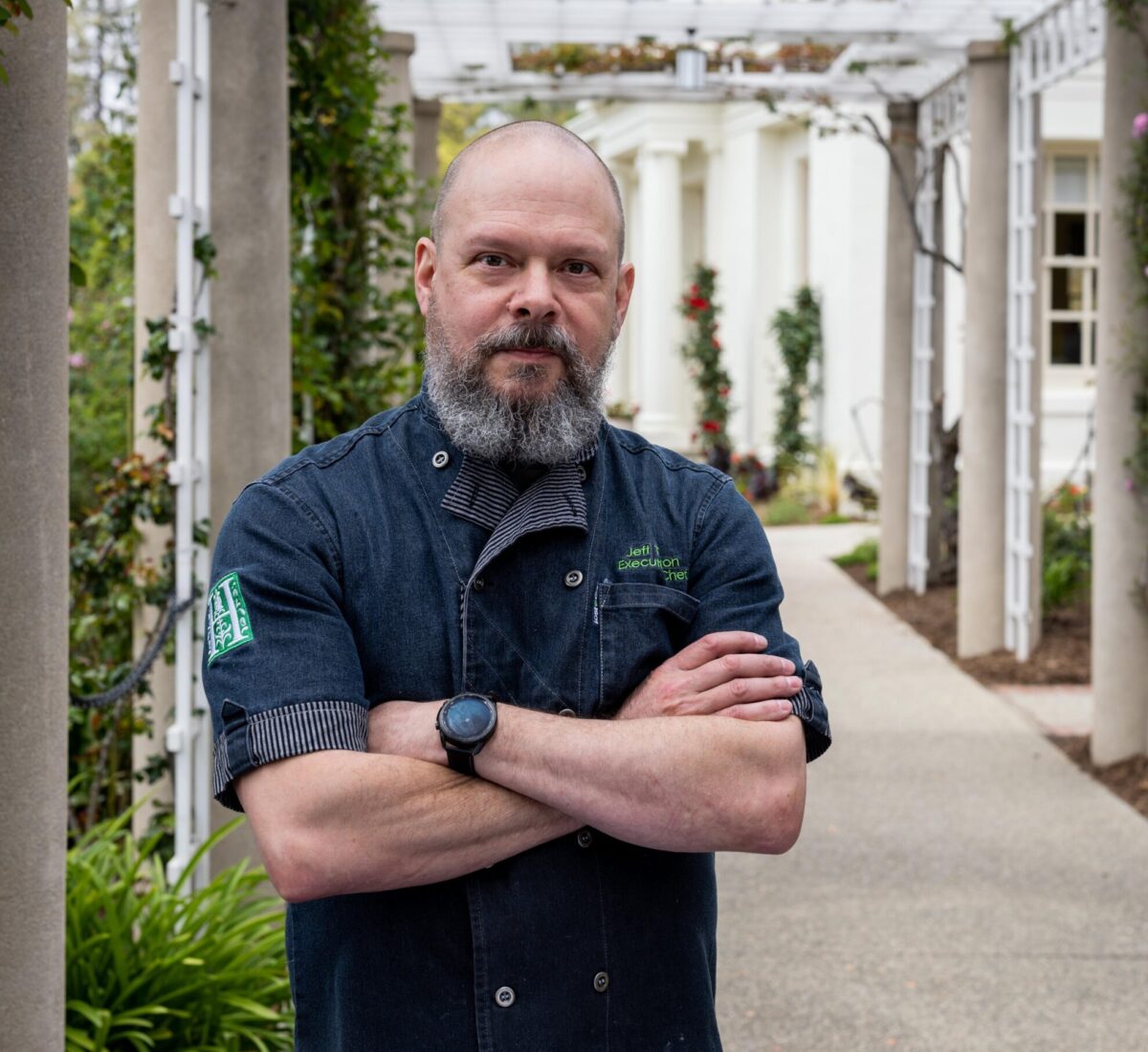 A man in a short-sleeved chef coat stands in front of a long portico