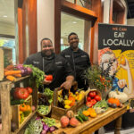 A man in a hat and a woman in a headscarf stand at a Farm to Table display.