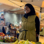 A young Black woman in a black headscarf stands behind a table full of produce.