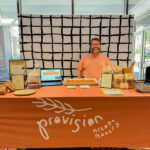 A man with a beard in a light orange shift stands at a display table with an orange cloth that says Provisions Bread and Bakery