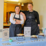 Two women wearing blue aprons stand behind a table with samples of baked goods.