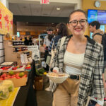 A young women wearing round glasses and a plaid shirt smiles with a bowl of food.