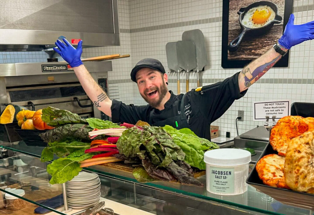 A man in a flat cap and a chefs' coat spreads his arms across a display of produce. 