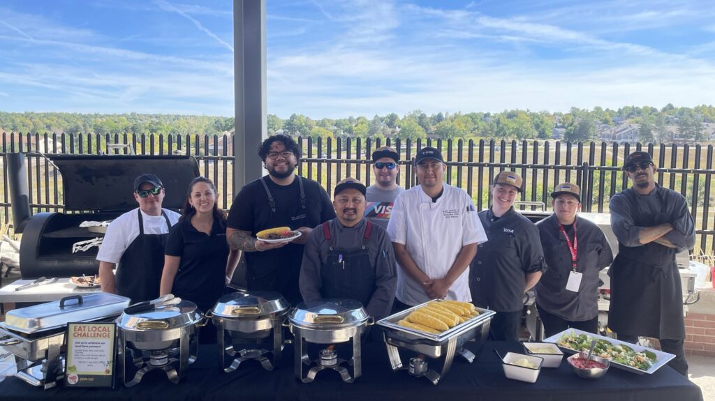 A group of culinary professionals stand together behind a catering setup with blue sky behind them. 