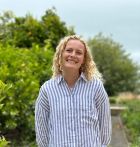 A young woman with curly blonde hair, wearing a blue and white striped buttondown shirt stands smiling in front of a stand of trees.