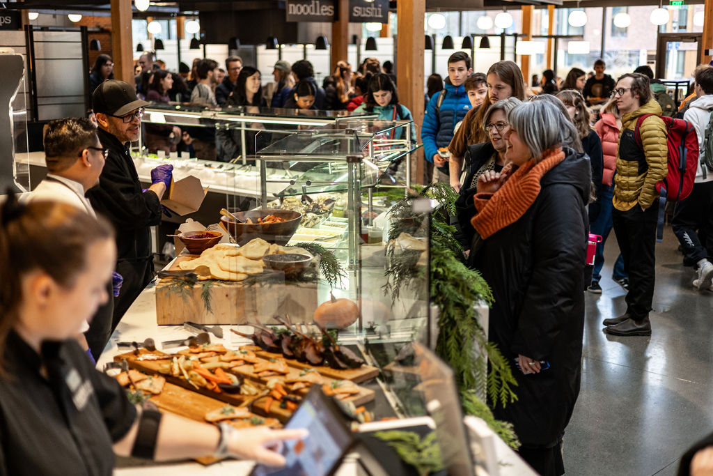 A crowd of people line up to be served food on platters by culinary workers.