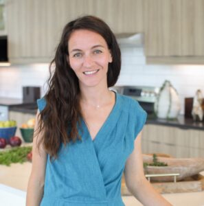 A woman with long brown hair wearing a blue dress is standing and smiling in a kitchen.