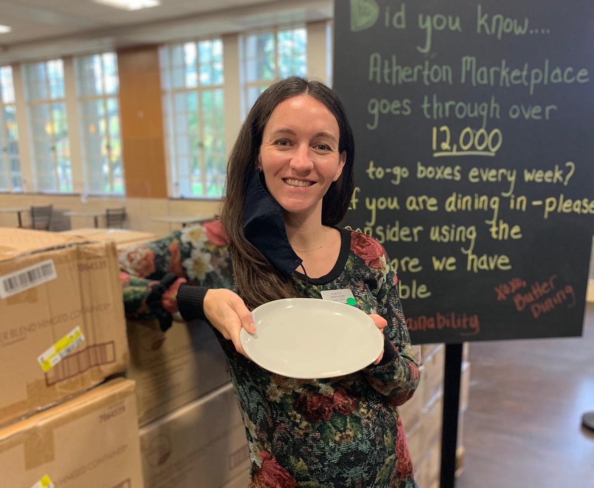 A woman holds a plate in front of a pile of boxes and a sign about to-go container waste