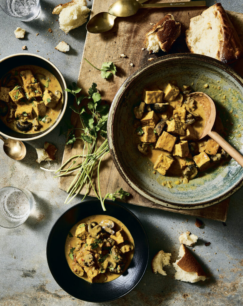 An overhead photograph of dishes of tofu-mushroom curry on a grey background with ingredients cattered around. 