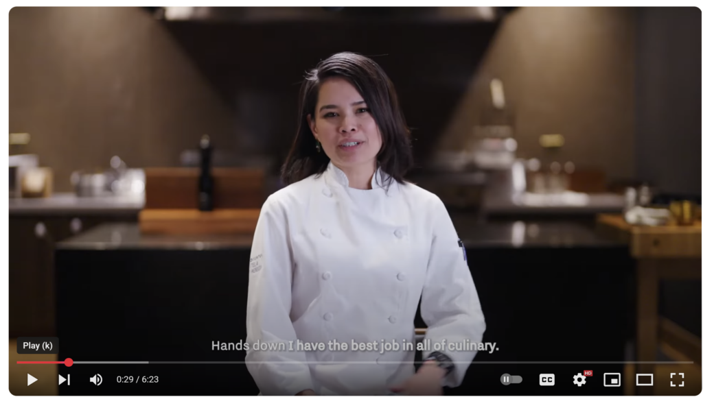 A woman chef in her whites sits in front of a warm-looing counter and speaks to the camera. 