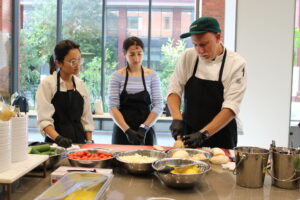 A man wearing culinary whites, an apron, and a baseball hat demonstrates knife skills to two women in a kitchen.