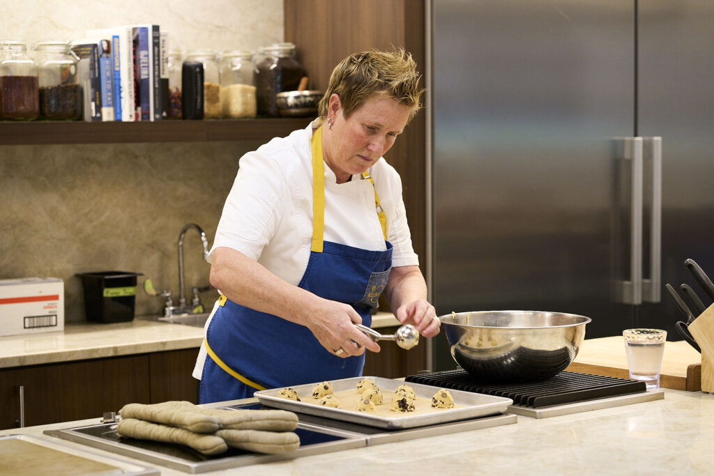 A woman in culinary whotes and an apron scoops chocolate chip cookies onto a sheet pan in a professional kitchen.