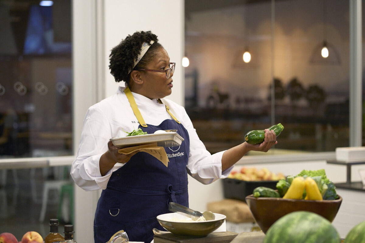 A woman chef stands in front of a group in a teaching kitchen setting. She's holding a zucchini.