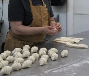 A person in a brown apron shapes croissants at a baking bench. 