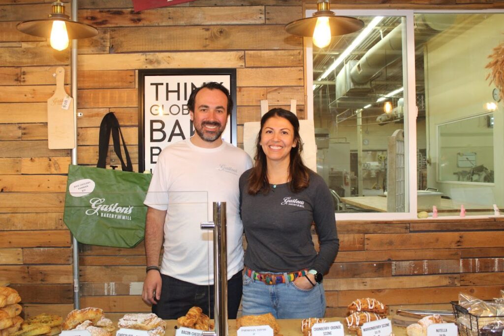 A man and woman stand together behind a display of baked goods. There is a window behind them looking into a baking operation. 