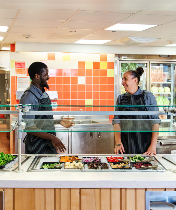 Two chefs, one man and one woman, chat while standing behind a salad bar.