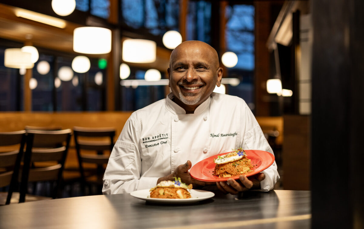 Chef Nimal Amarasinghe sits in his chef coat holding a plate of food in a dining room