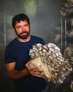 A dark-haired man with a beard holds a very large oyster mushoom