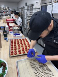 Pastry chefs work at their bench. In the foreground, a chef in a black cap does detail chocolate work. 