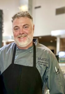 A chef in a dusty green chef's coat and a black apron stands smiling in a commercial cafe.