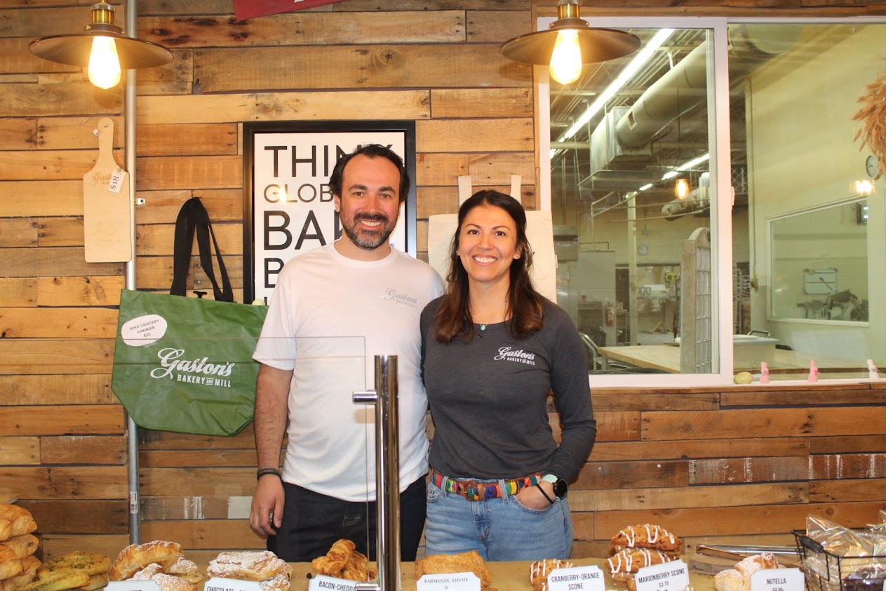 A man and woman who own a bakery stand behind a spread of their baked goods.
