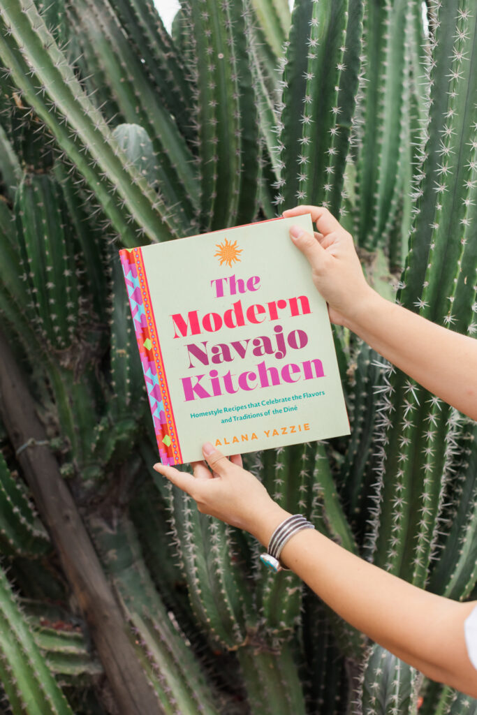 Someone holding a cookbook called The Modern Navajo Kitchen in front of a large cactus