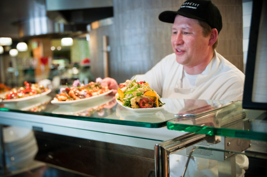 A chef in a white chef's coat and black cap passes food over a counter to a guest