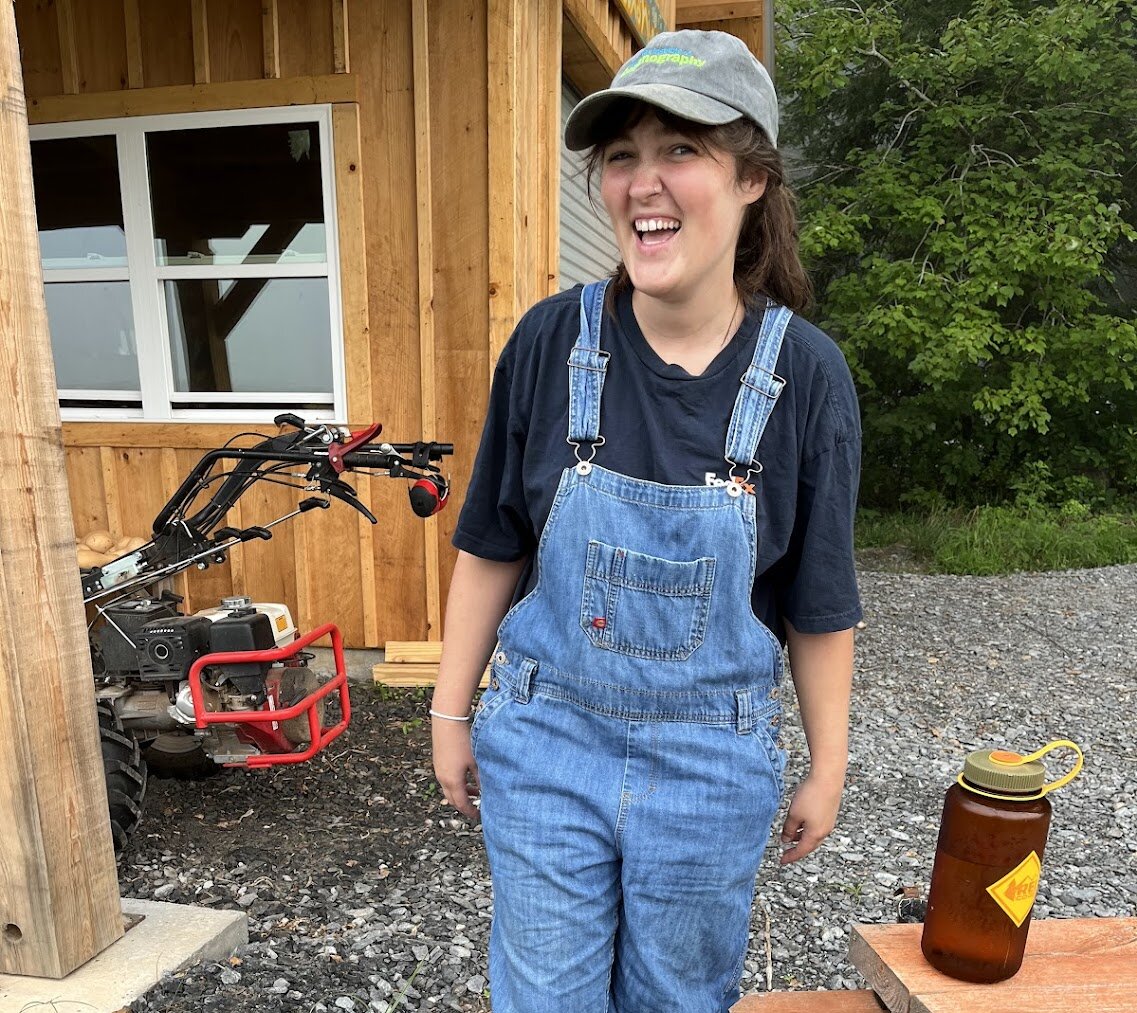 A young woman in a ball cap and coveralls smiles at the camera during a break from working on a farm