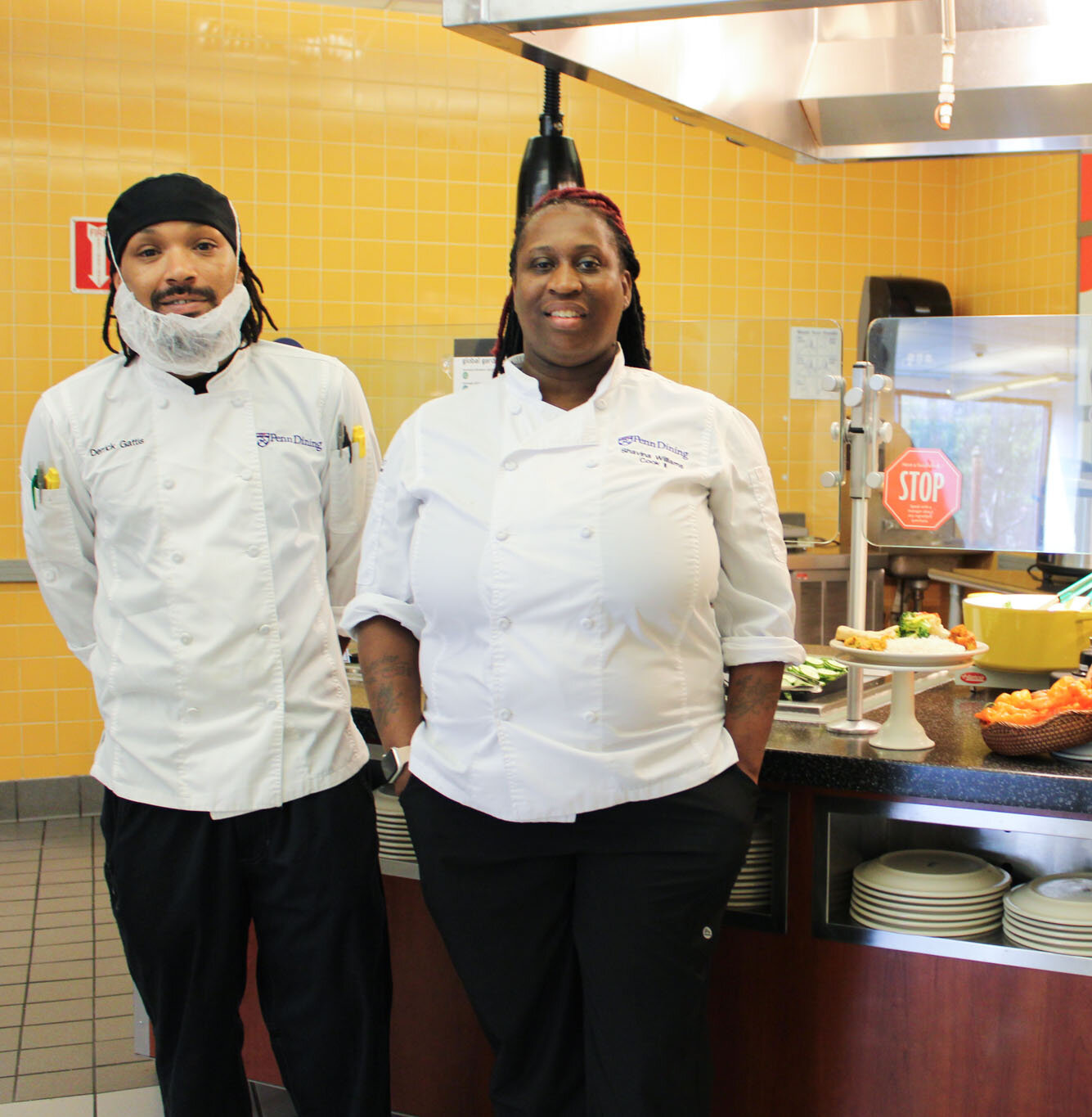Two culinary professionals in white chef coats stand in front of a dining station at Penn.