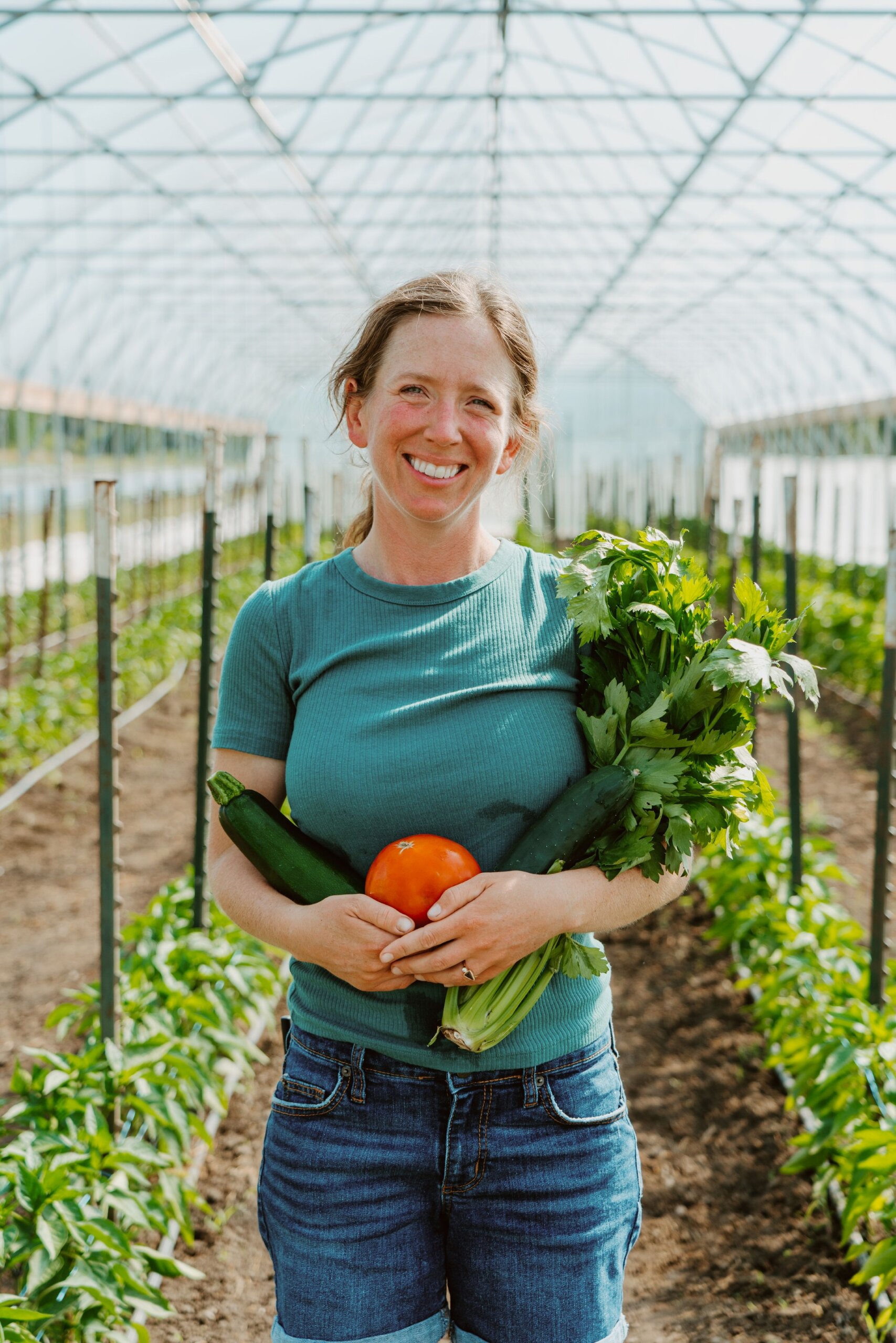 A young woman with a brown ponytail stands in a greenhouse with produce growing behind her. She is holding a tomato, a cucumber, and some leafy greens.