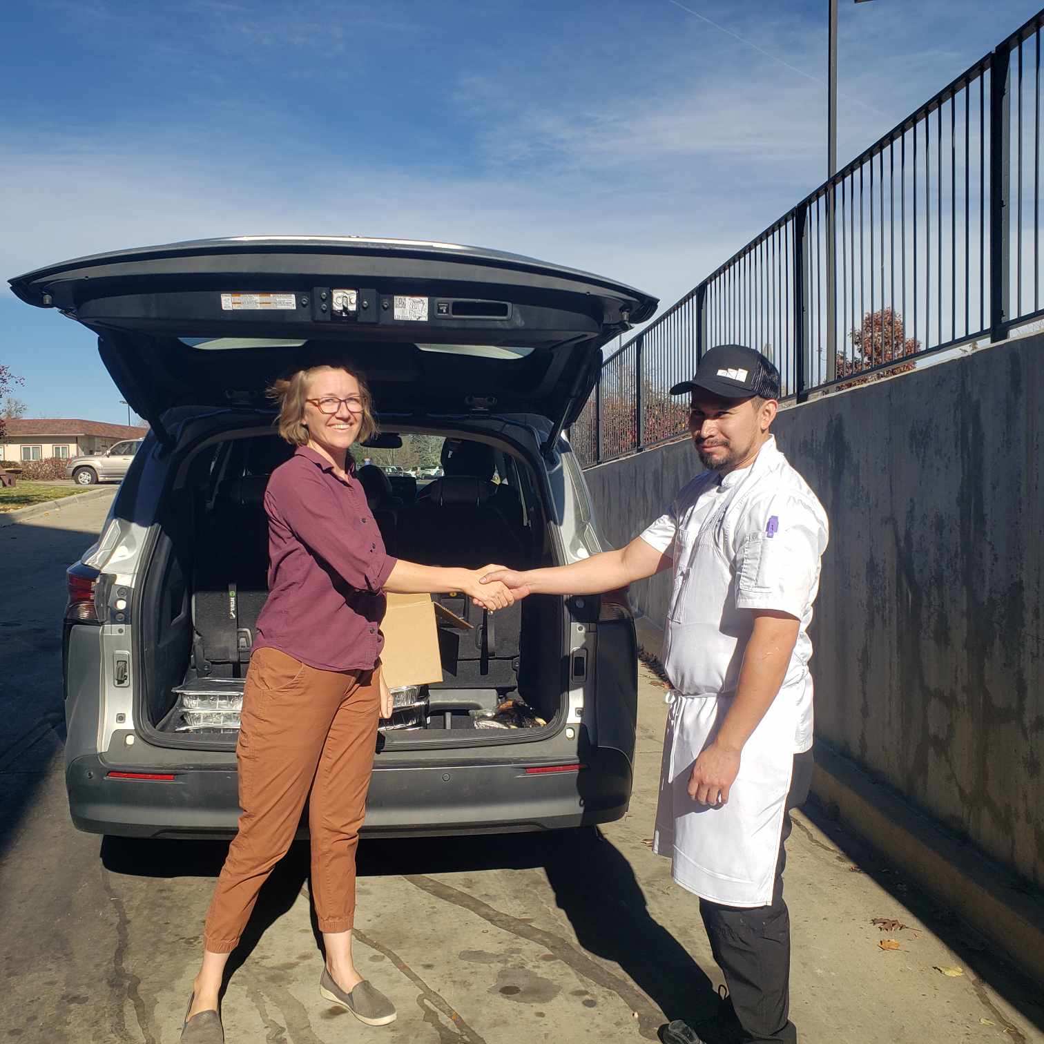 A man and a woman shake hands behind the hatch of a car filled with recovered food