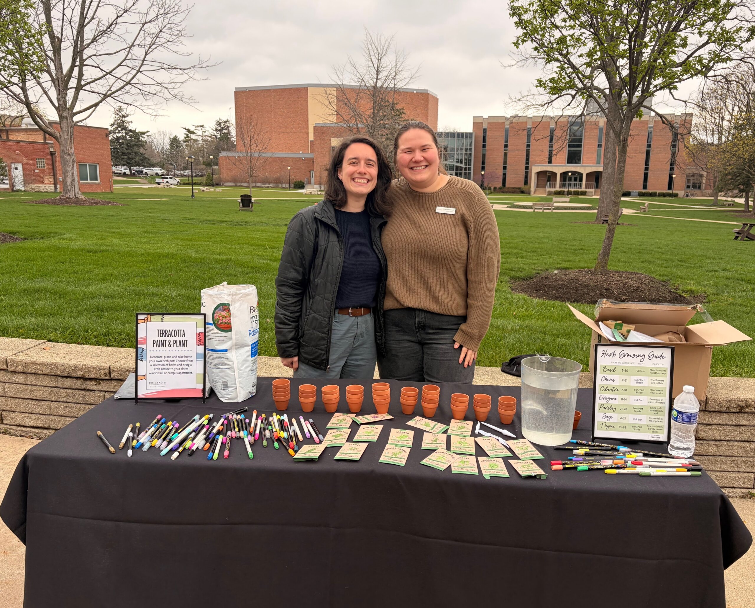 Two young women stand together at an outdoor activity table on a college campus.