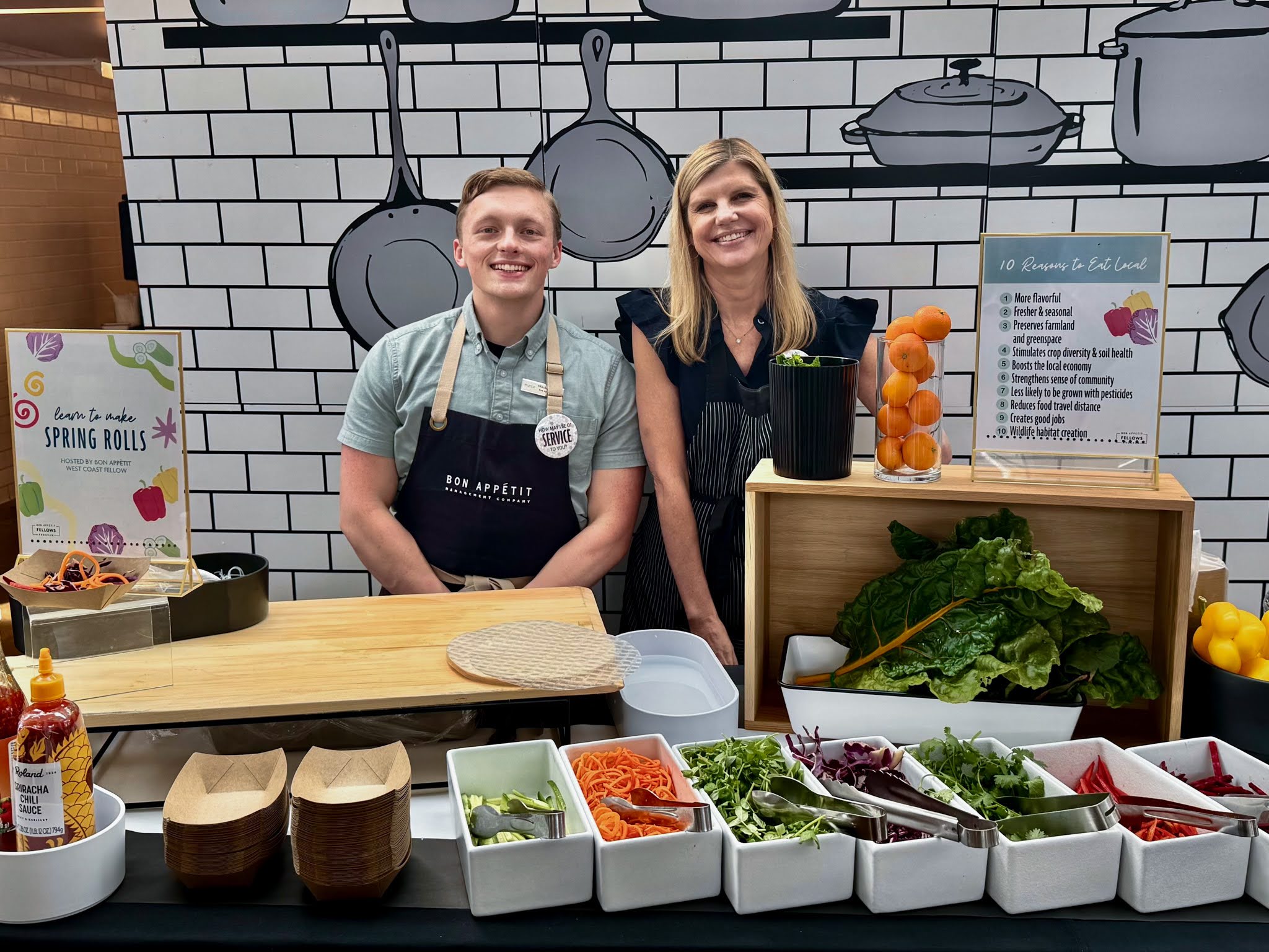 A man and a woman, both Bon Appetit Management Company employees, stand together behind a Local Food display