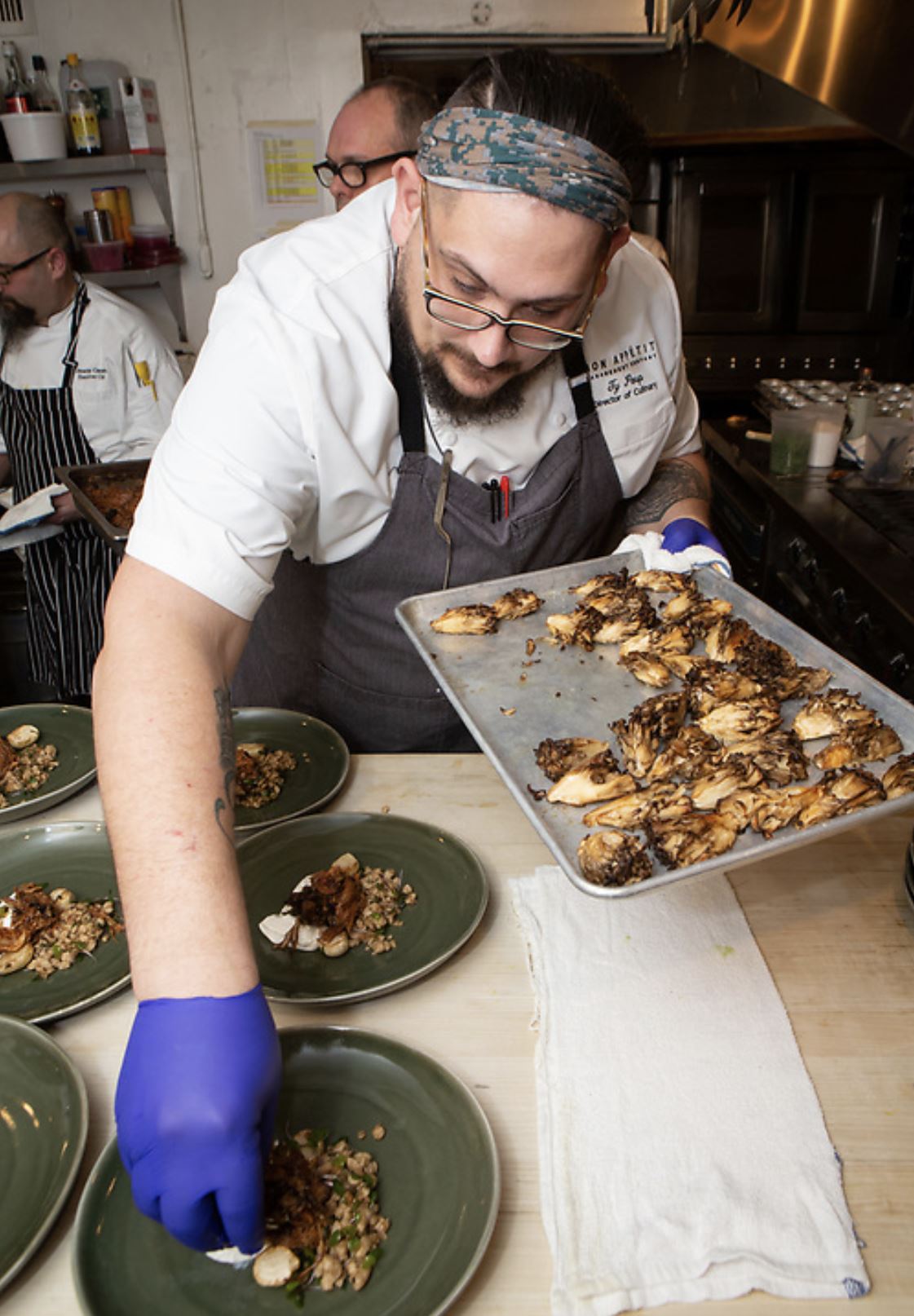 A chef in blue gloves and a headscarf places roasted vegetables onto a row of plates.