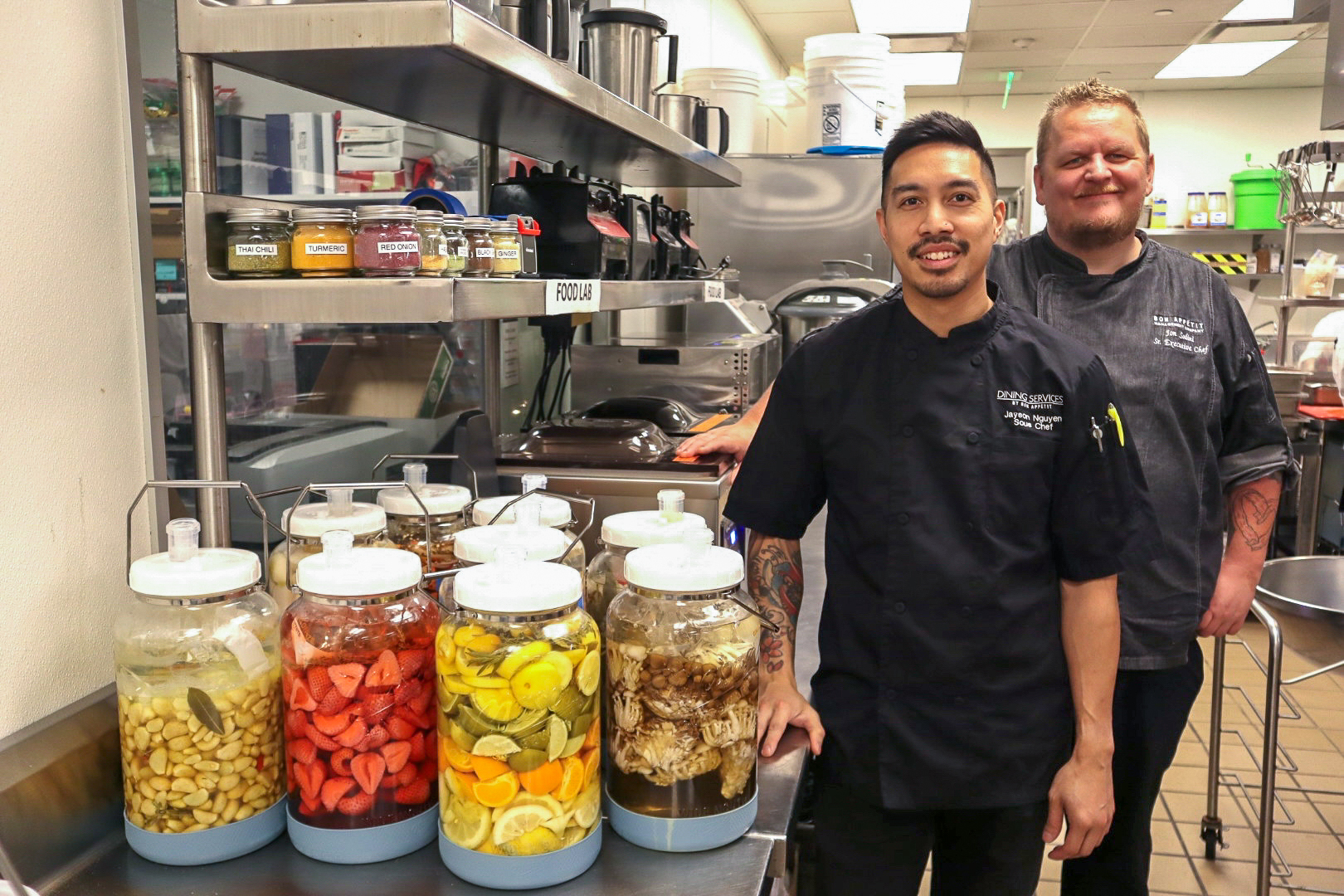 Two chefs stand in a commercial kitchen with their fermented creations lined up in front of them.