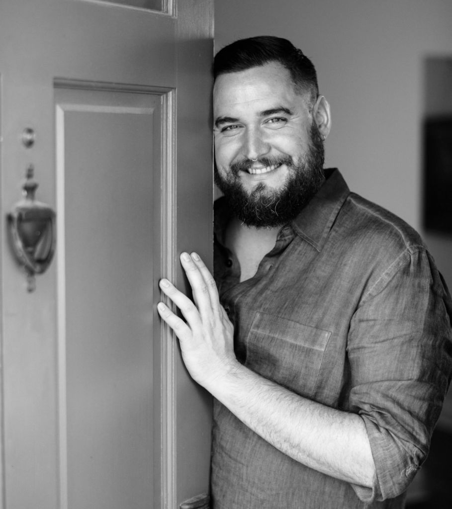 Black ad white photo of a smiling bearded man at his front door